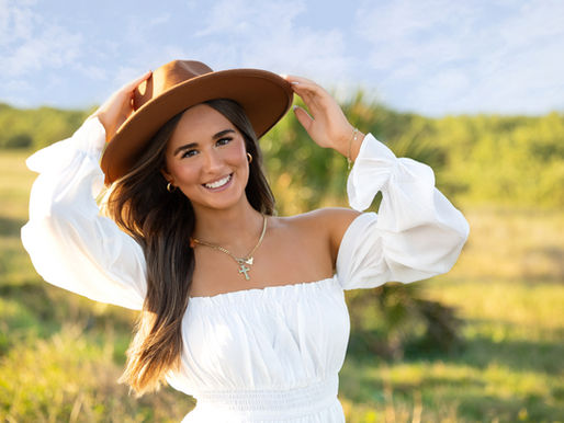 High school senior girl in white dress and wide-brimmed hat smiling during golden hour senior session in St. Augustine, Florida