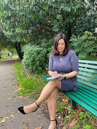 Woman in a gray dress sits on a park bench, writing in a notebook.