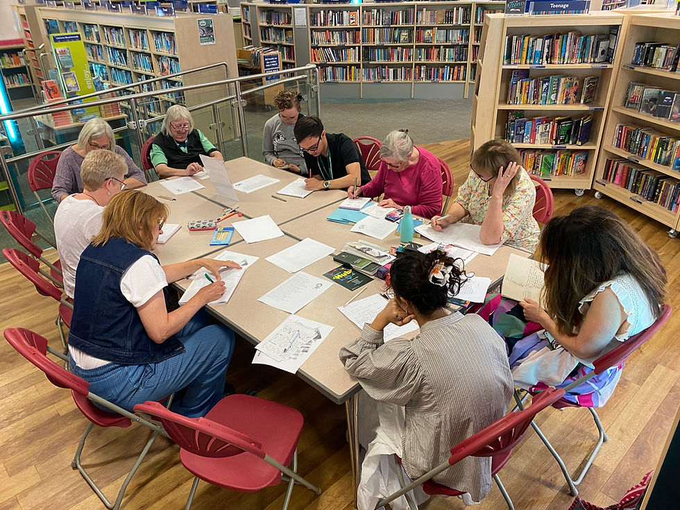 A group of people sit around a table writing in a library with bookshelves in the background. Red chairs and wood flooring are visible.