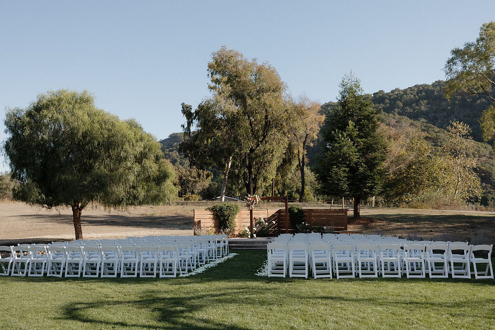 Outdoor wedding setup with rows of white chairs on a grassy lawn, floral aisle, trees and hills under a clear sky at Tar Creek Ranch by SLO Wedding Photographer