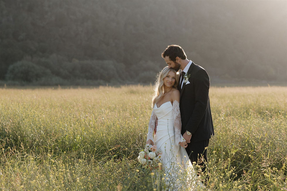 Bride and groom stand in a sunlit meadow, he kissing her head as she smiles in a white gown holding a bouquet.