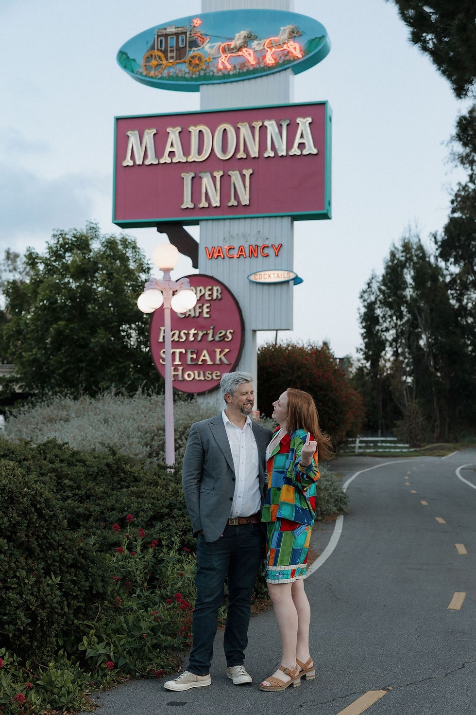 Smiling couple stands by the Madonna Inn sign with neon Vacancy and Cocktails, on a curving road at dusk.