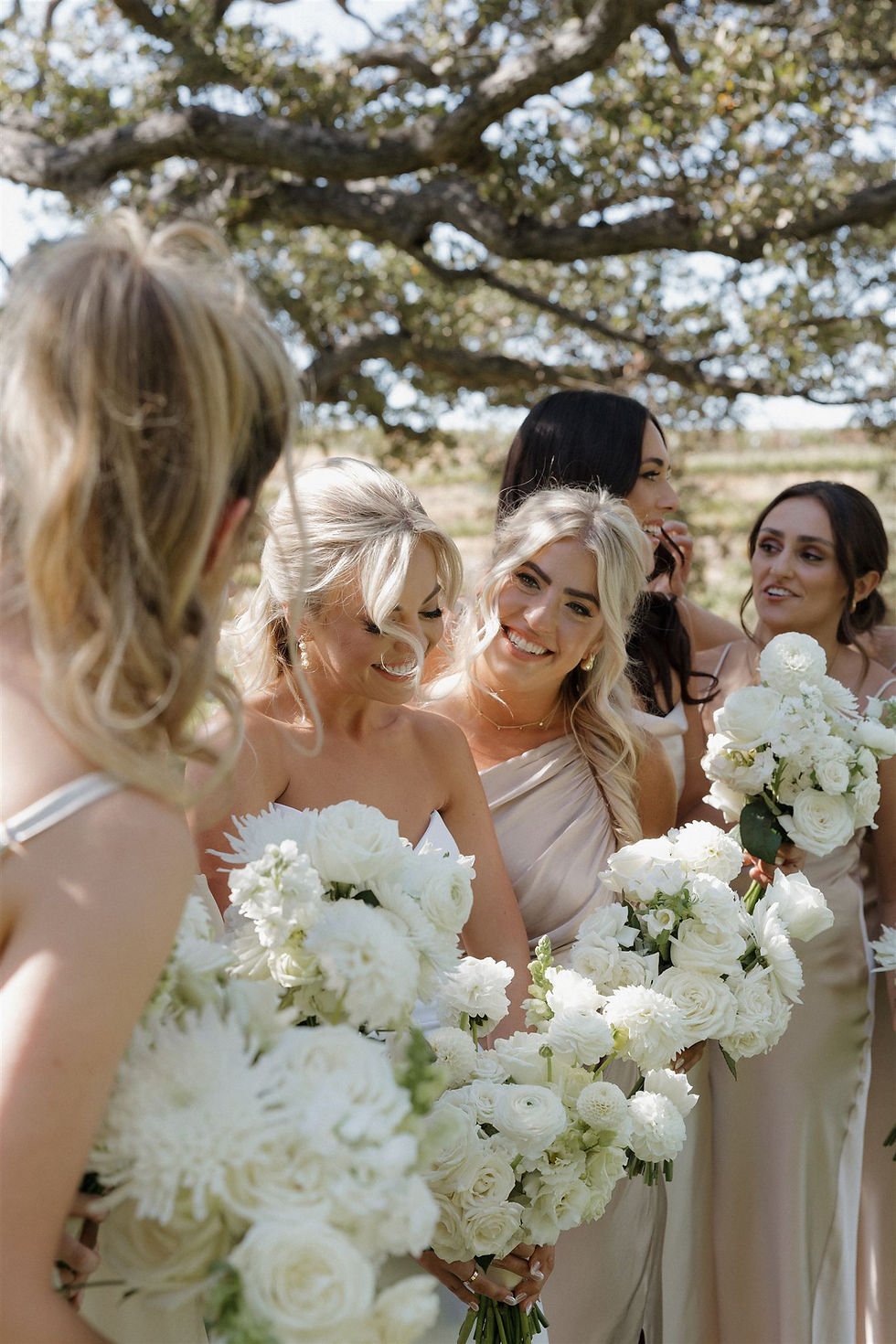 Bridesmaids in light dresses smile and hold white flower bouquets under a tree, creating a joyful and serene outdoor scene during First Look with Bridesmaids in a Paso Robles wedding