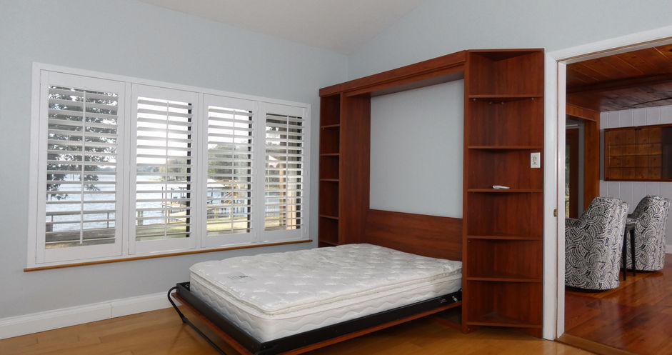 Bedroom with a bed, wooden shelves, and window shutters overlooking the lake.