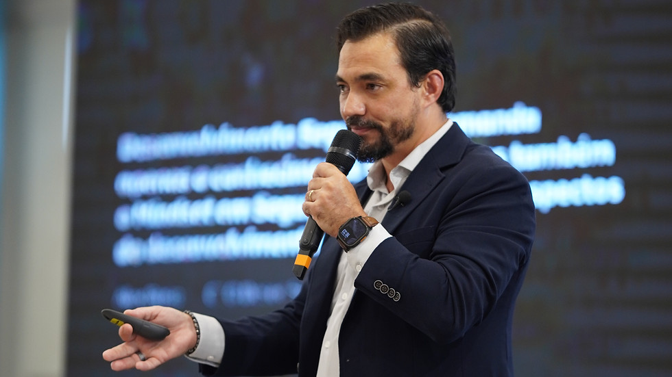 Foto em formato retrato de um palestrante em evento corporativo, com fundo desfocado e cores sóbrias