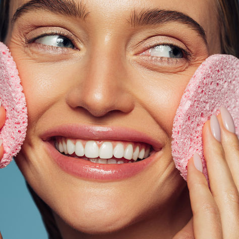 Smiling woman double cleansing her face with two pink facial sponges
