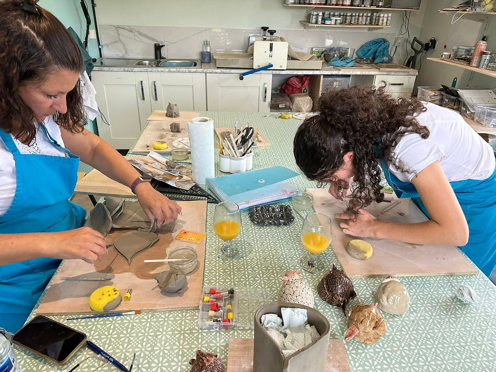 Two women in blue aprons crafting leaf-shaped clay pieces at a table filled with pottery tools