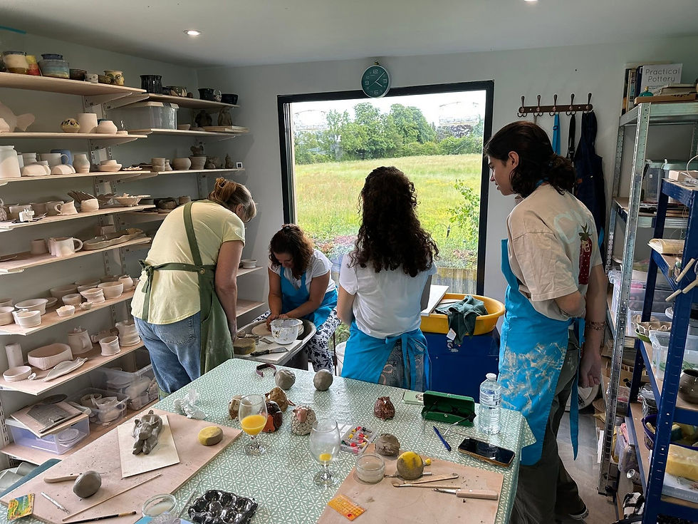 Woman sitting at a craft studio table detailing small ceramic sculptures surrounded by pottery glazes, paintbrushes, and clay vessels