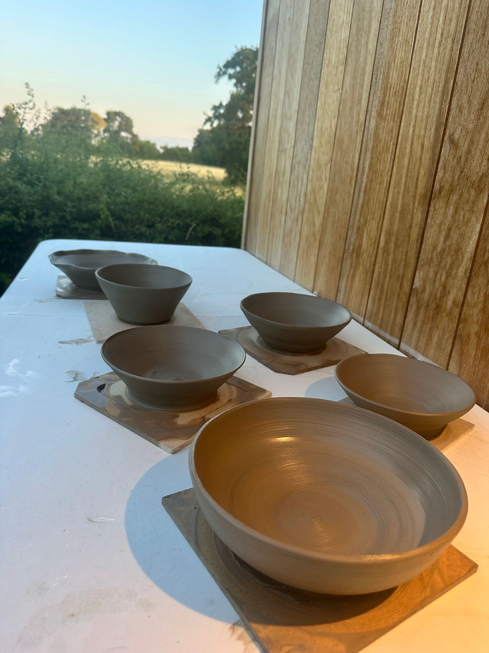 Row of freshly thrown raw clay bowls drying on wooden bats outdoors on a white table