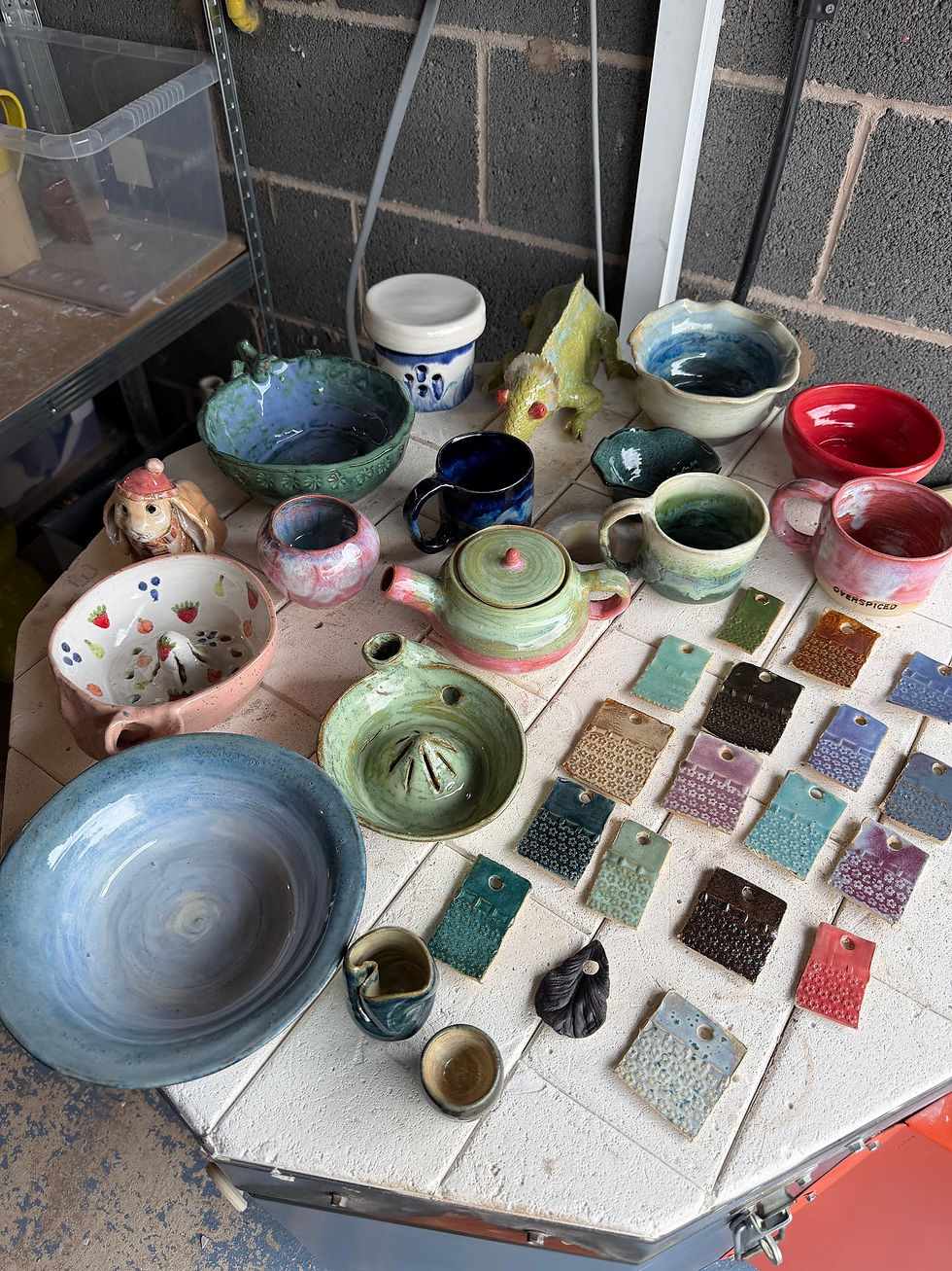 Collection of colourful glazed pottery including teapots, bowls, mugs, and ceramic glaze test tiles arranged on a table