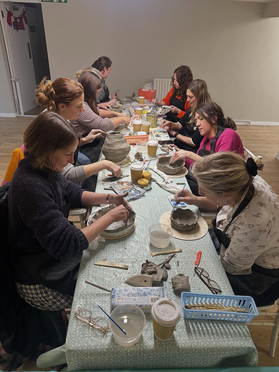 Group of people sitting along a long table during an indoor pottery workshop sculpting clay pots together