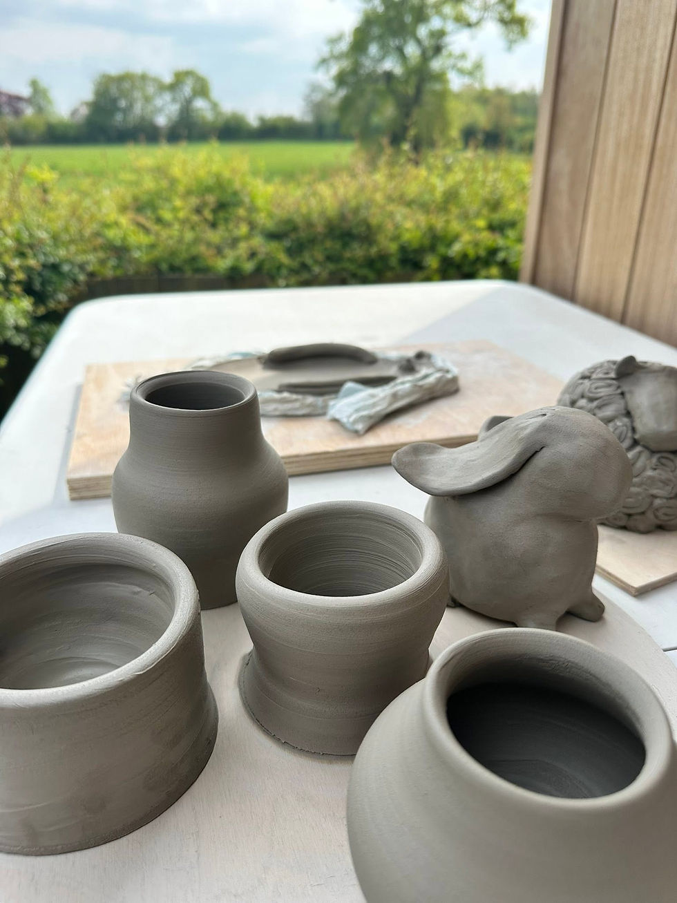 Hand-thrown raw clay pots and a sculpted rabbit figure sitting on an outdoor workbench