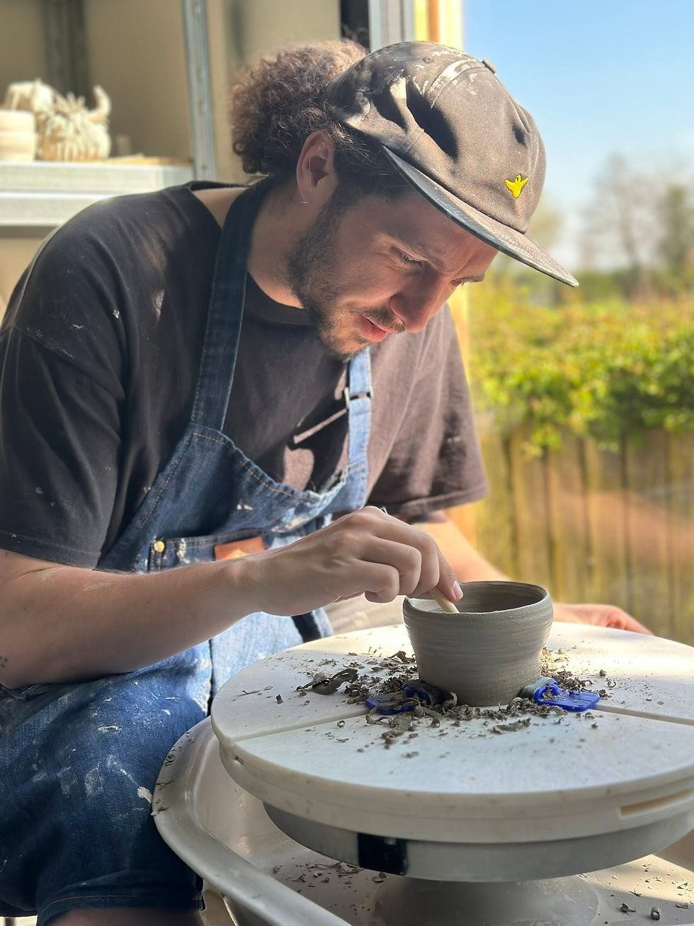 Man in a cap and denim dungarees detailing a small clay pot on a pottery wheel