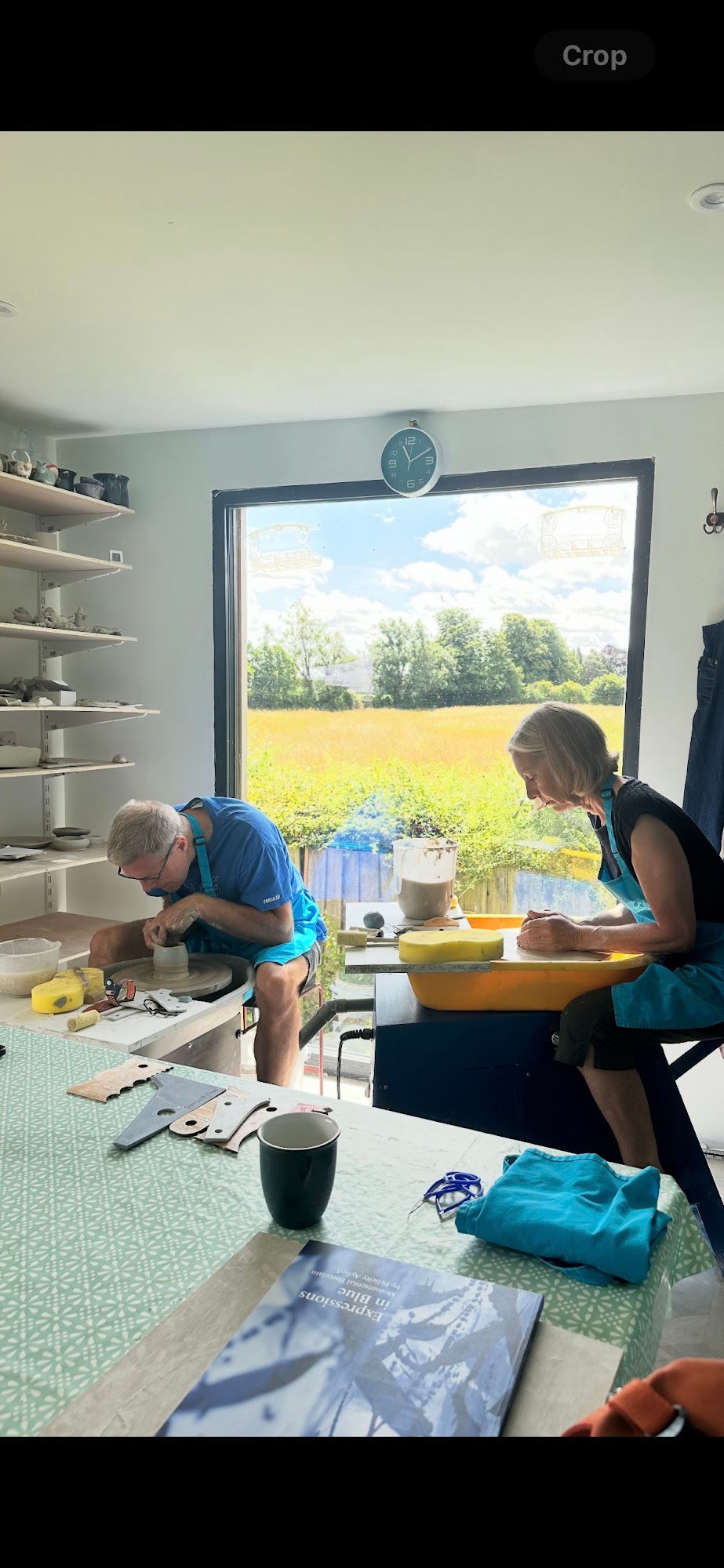 Man and woman sitting at potter's wheels throwing clay in front of a window in a countryside studio
