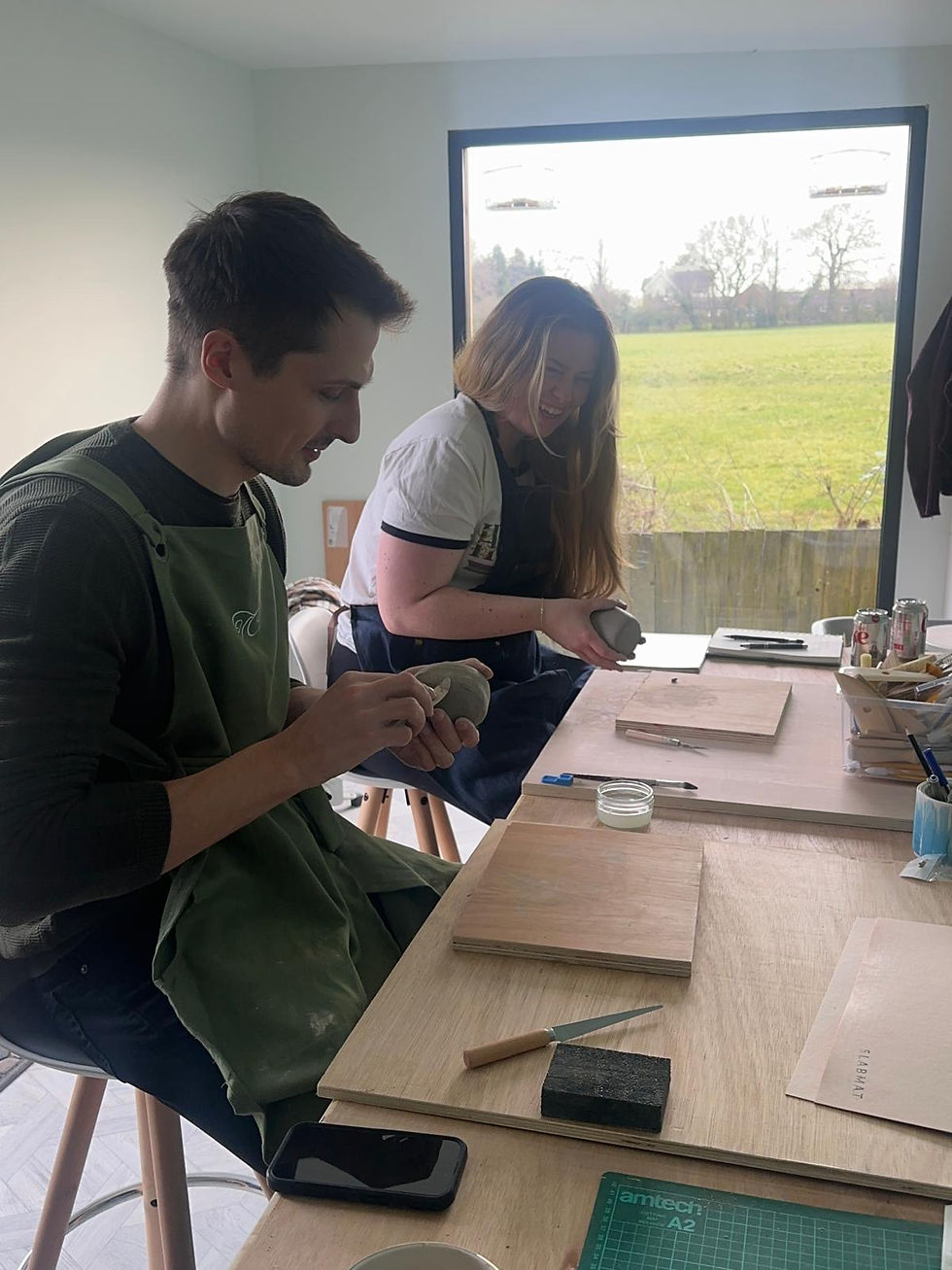 Man and woman smiling while hand-building ceramic pieces together at a pottery workbench in a bright studio
