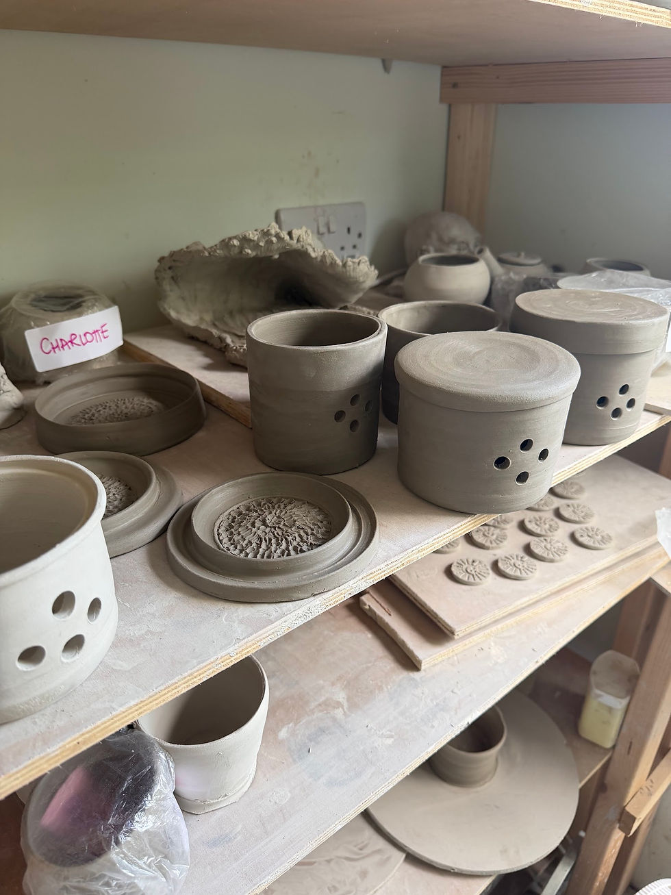 Unfired clay pots, garlic keepers with holes, and stamped clay discs drying on wooden shelves in a pottery studio