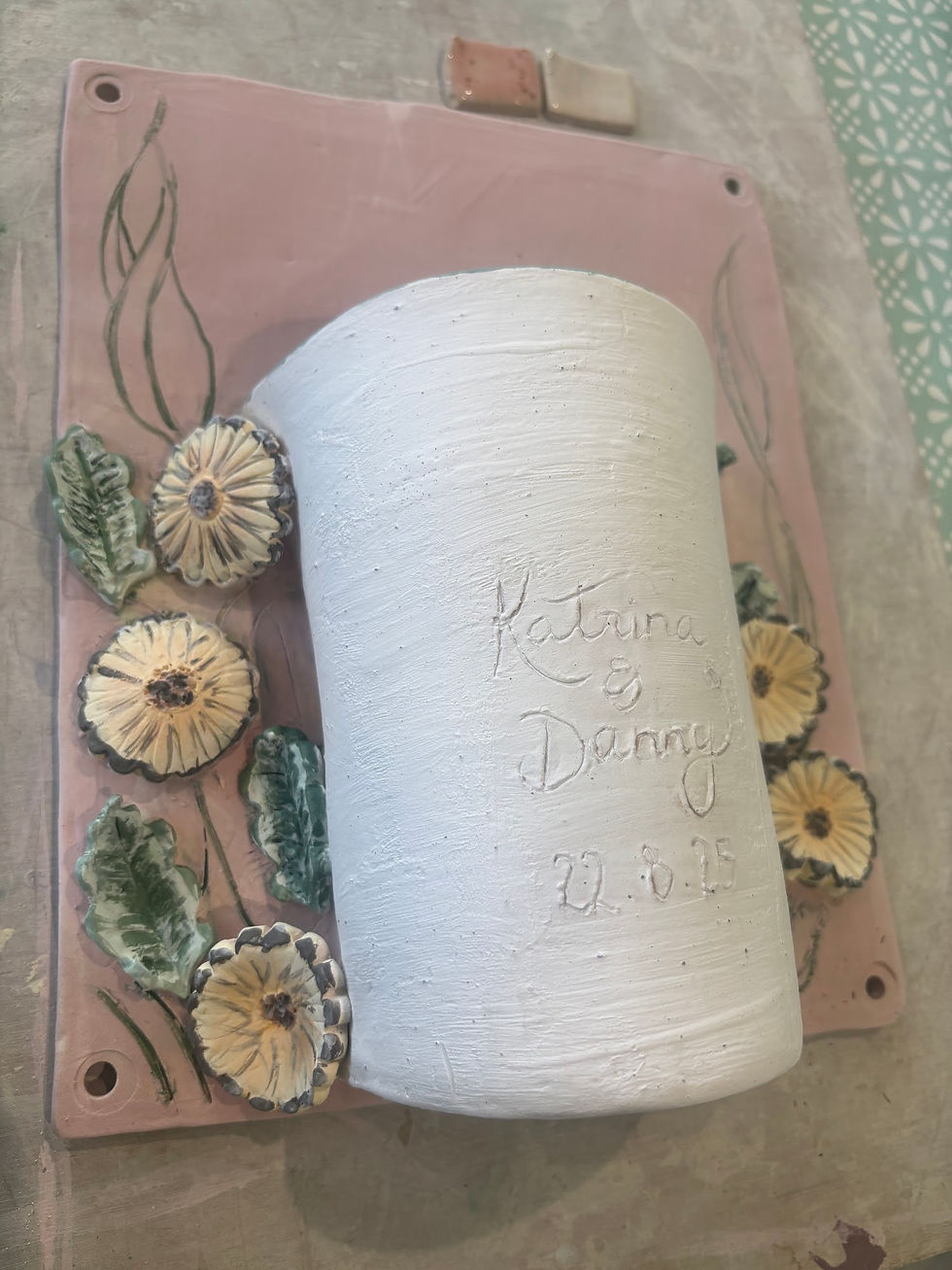Personalised ceramic wall plaque featuring handcrafted clay flowers and inscribed names lying on a workbench
