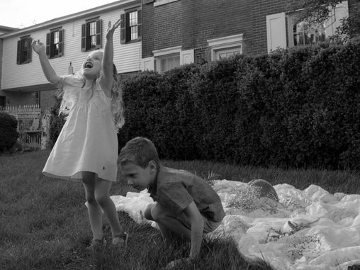 Young girl joyfully raises arms, boy crouches on grass beside blanket in a residential backyard. Brick houses and hedges in background. Black and white.