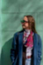 Woman in sunglasses and a blue coat stands against a green wall, wearing a colorful scarf. She's looking to the side, exuding a relaxed mood.