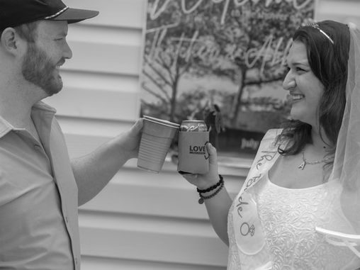 Man and woman in festive attire toast with drinks, smiling. "Love Muskegon" visible on cup. "Welcome" sign in background. Joyful mood.