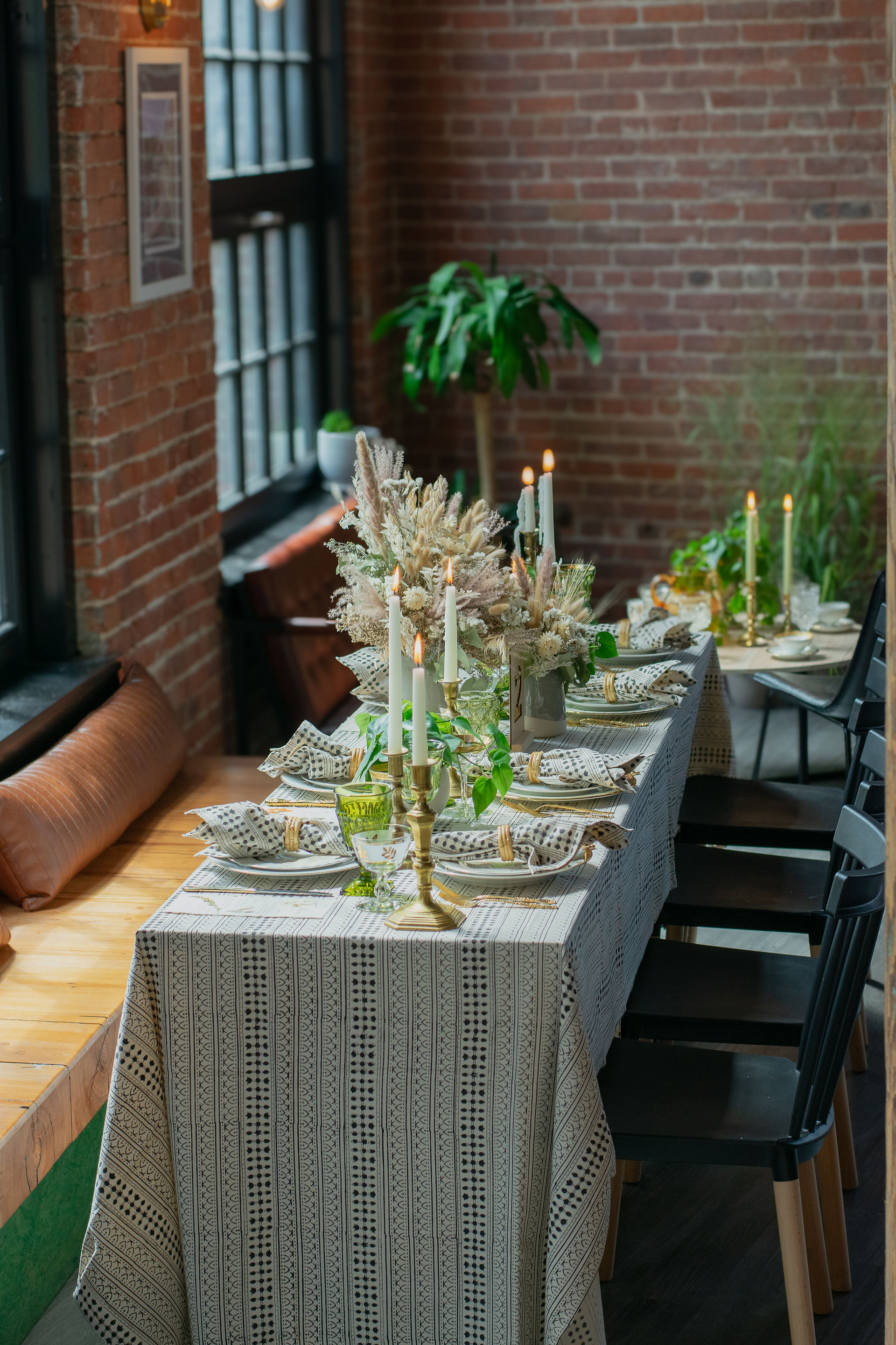 Elegant dining table set with patterned cloth, candles, green glasses, and dried flowers. Brick wall and indoor plant in background. Cozy ambiance.