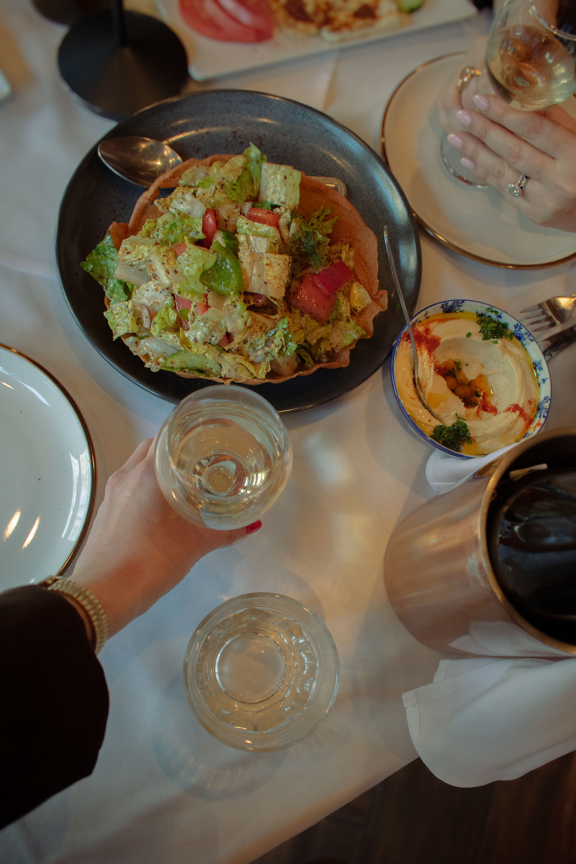 Woman with long hair holding a wine glass, sitting at a restaurant table with dishes and a wine bottle. Dim, cozy ambiance.