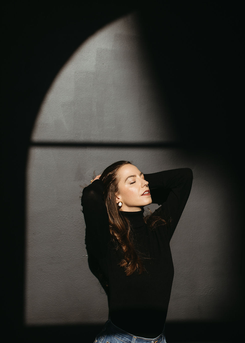 Woman in black turtleneck and jeans poses with hands on head, eyes closed, in dramatic sunlight against a dark, textured backdrop.