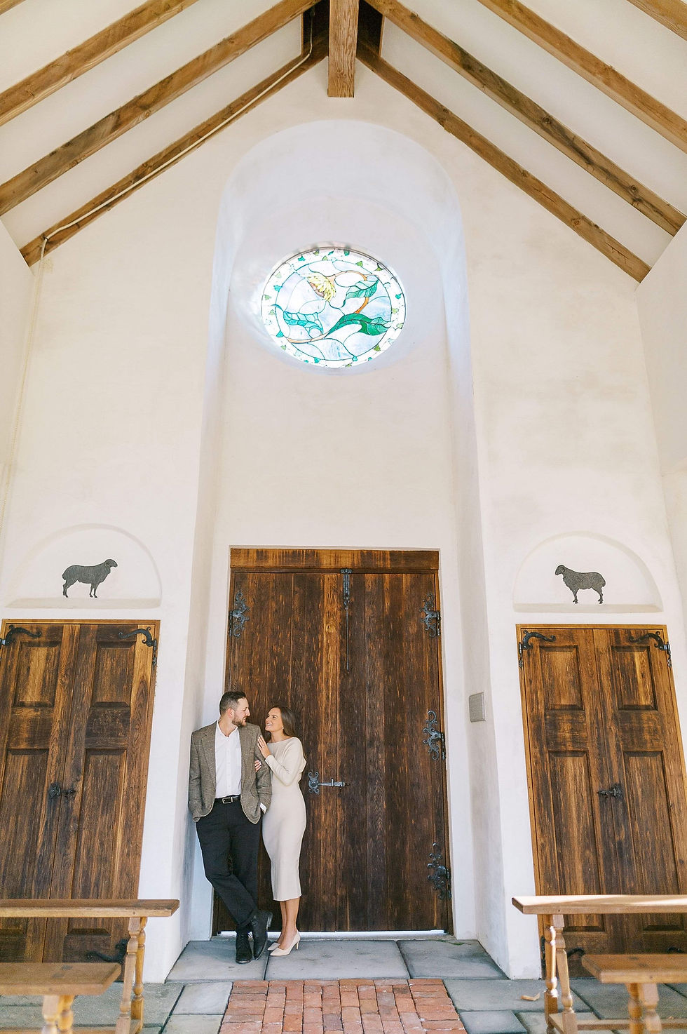 Man in sports coat with black pants and black hair embracing woman in long cream colored sweater dress in a small open outside chapel in the woods.