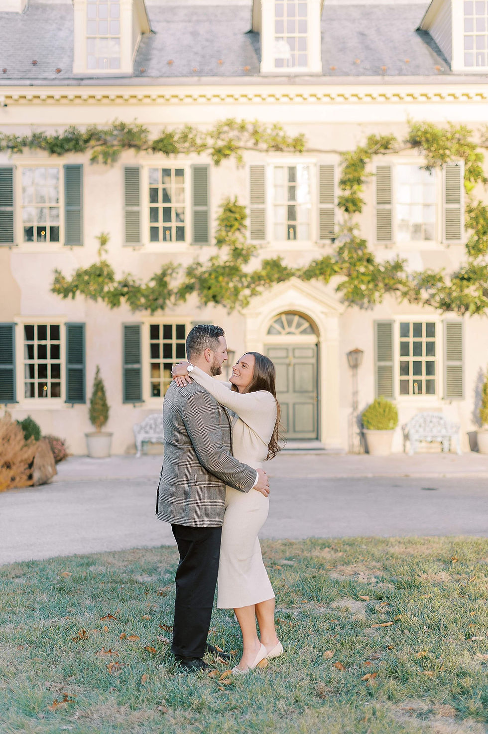 Engagement Session at Hagley Museum in Wilmington, Delaware, man in sport coat and black pants embracing in on woman with dark brown hair in long cream colored sweater dress and matching heals in front of the big house on the lawn in fall.