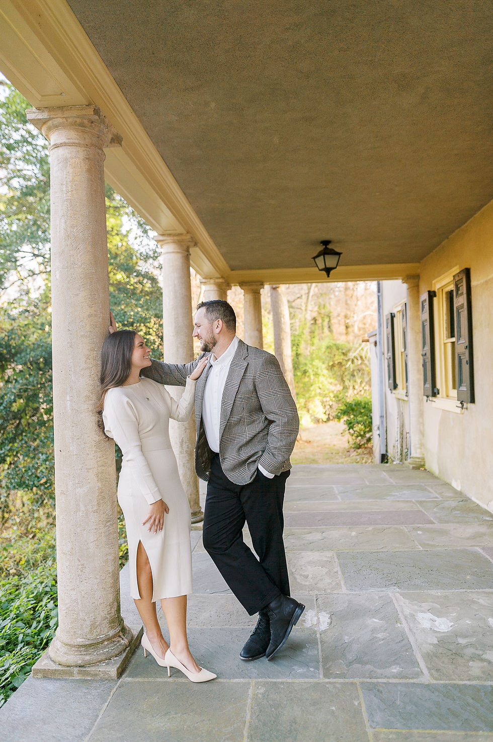 Engagement Session at Hagley Museum in Wilmington, Delaware, man in sport coat and black pants leaning in on woman with dark brown hair in long cream colored sweater dress and matching heals
