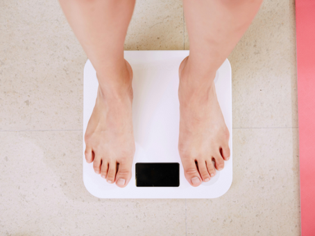 Top-down view of a person standing barefoot on a digital bathroom scale, symbolizing weight monitoring, health tracking, or weight loss journey. A pink yoga mat is partially visible to the side, suggesting a fitness or wellness context.