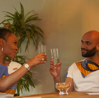 Een man en een vrouw zitten aan een tafel en toosten met glazen water. Beiden dragen bijpassende witte shirts met kleurrijke Afrikaanse waxprints. De setting is warm en ontspannen, met een plant op de achtergrond die een vleugje groen toevoegt.