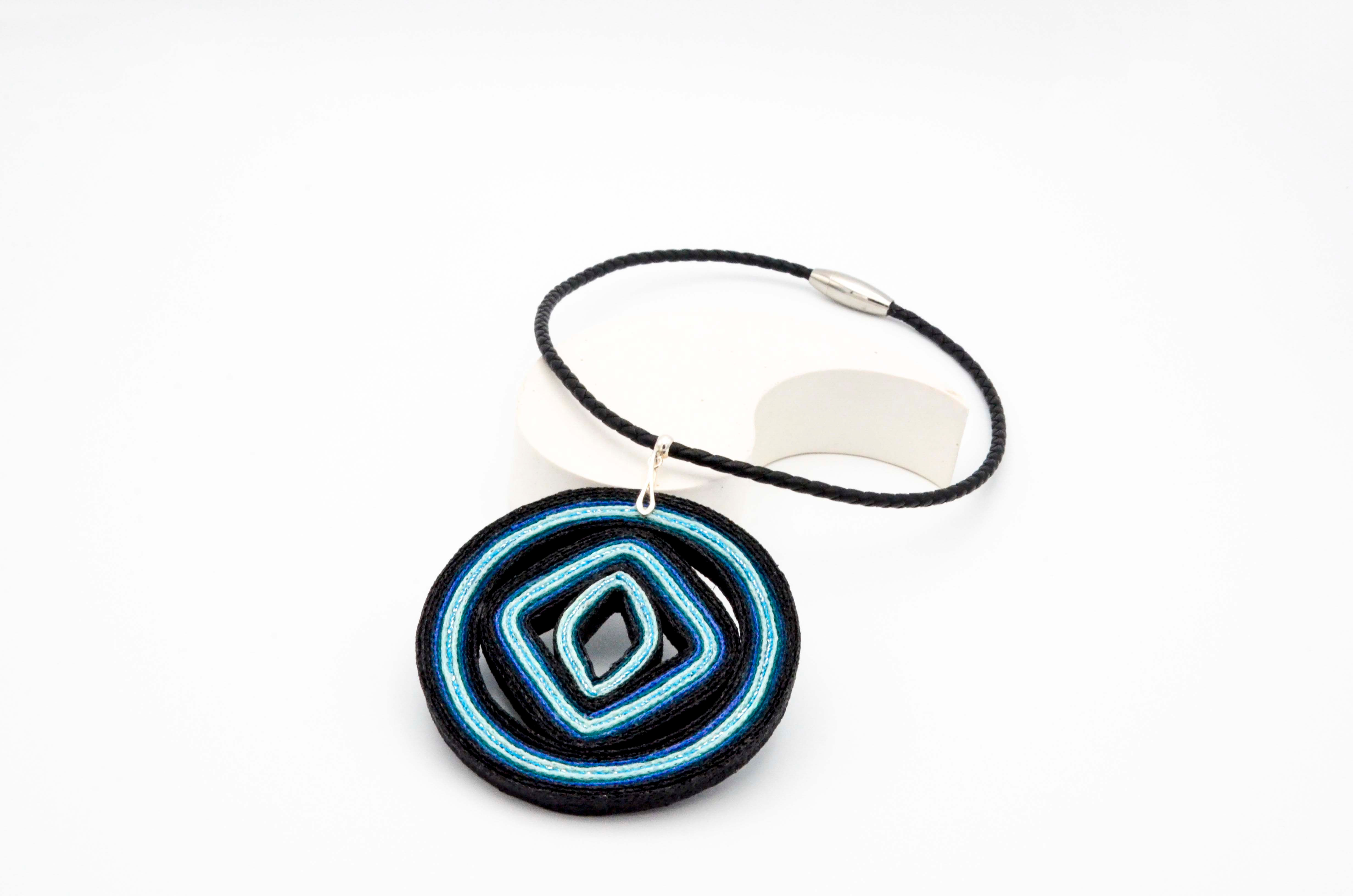 Colgante textil en tonos azules y negro con cordón de cuero trenzado color negro y cierre magnético