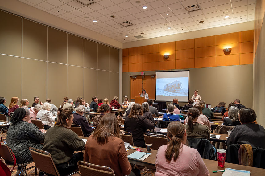Room of people sitting at tables facing a screen and two presenters.