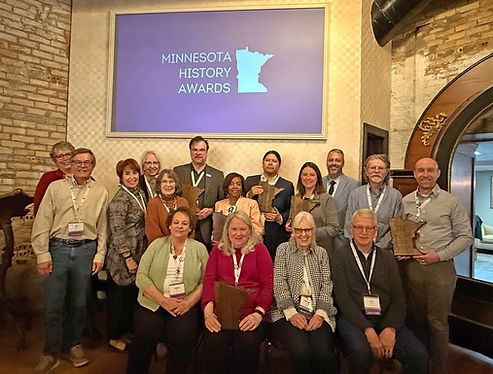 Sixteen people, some holding plaques, sit and stand in front of a screen projecting Minnesota History Awards.