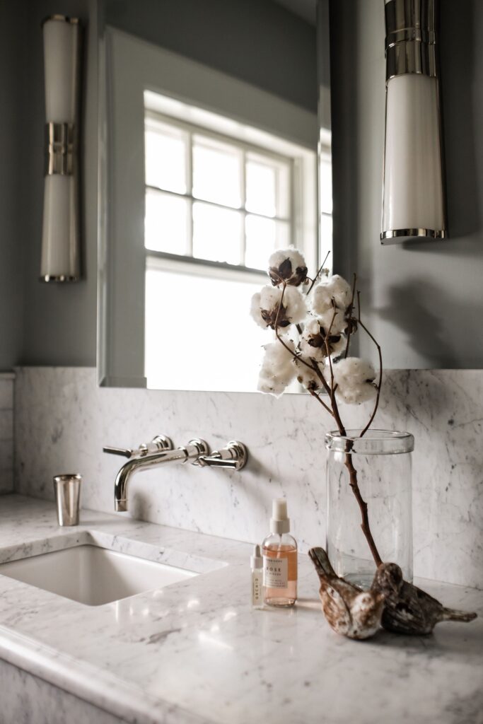 Marble bathroom counter with cotton flowers in a glass vase, skincare bottles, and a metal cup. Soft light from a window creates a calm vibe.