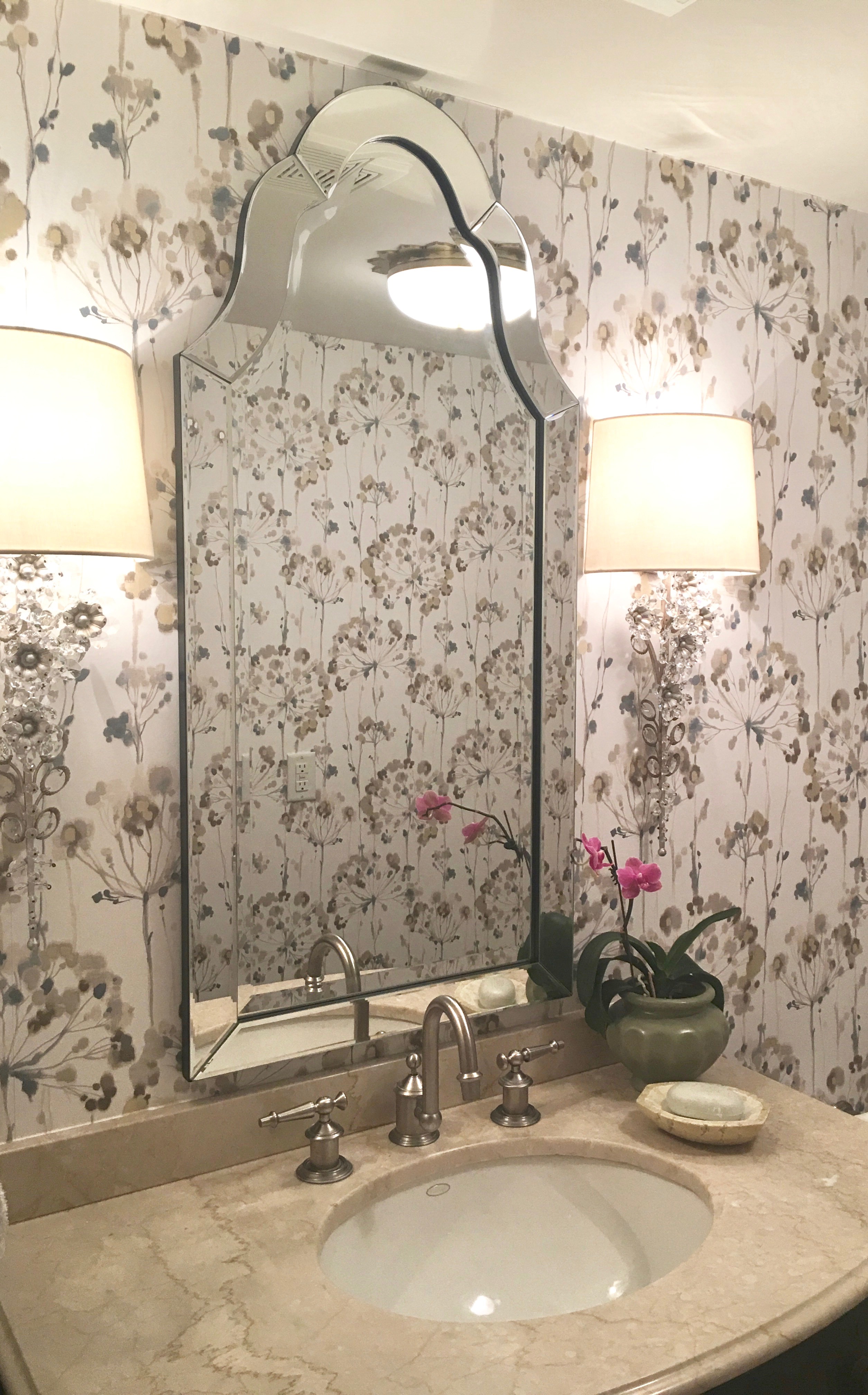Elegant bathroom with floral wallpaper, ornate mirror, and two lamps. Marble countertop with sink, soap dish, and potted orchid.