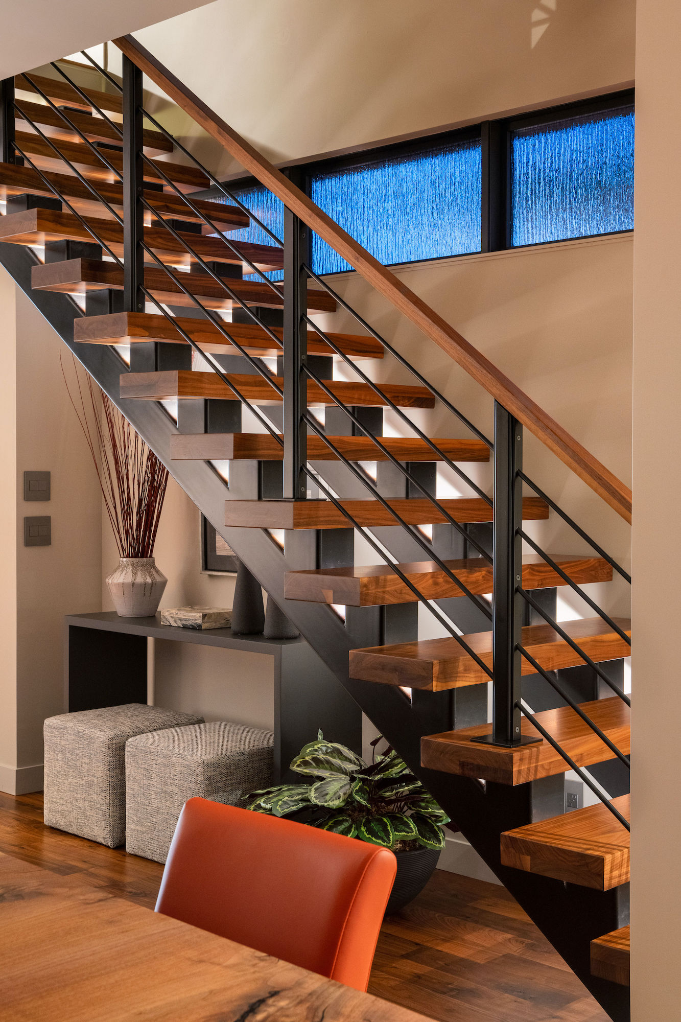 Modern staircase with wooden steps, black railings, and blue-tinted windows. Gray stools and a red chair add color to the serene setting.