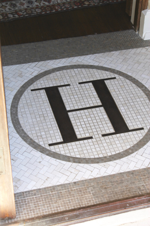 Mosaic floor with a large black letter "H" inside a circle against a white tile pattern. Wooden floor and rug visible in the background.