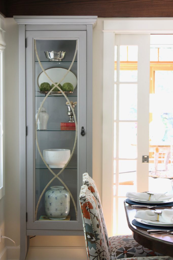 Gray cabinet with glass doors displays decorative bowls and a vase. Floral-patterned chairs and a set dining table in a bright room.