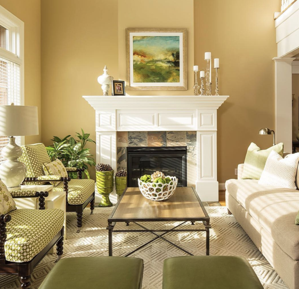 Cozy living room with a beige sofa, green chairs, and a white fireplace. Art above fireplace. Earthy tones and a plant add warmth.