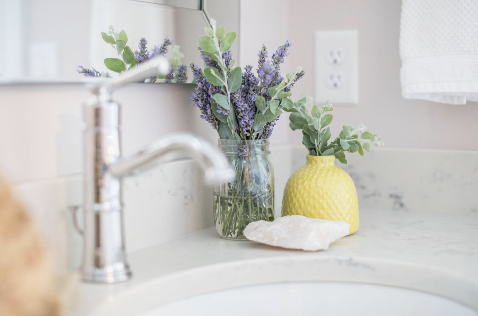 Purple lavender in a mason jar and green leaves in a yellow vase sit by a silver faucet on a marble bathroom counter. White towel nearby.