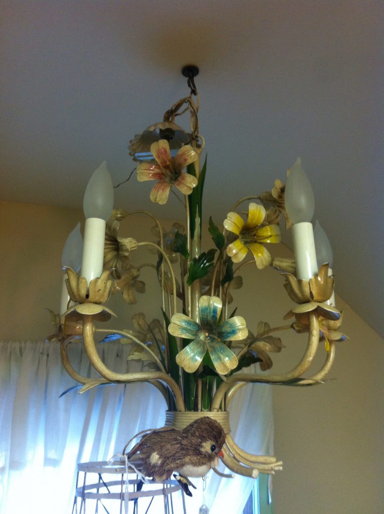 Floral chandelier with pastel flowers and candle-style bulbs hangs from ceiling; small decorative bird perched below against a beige wall.