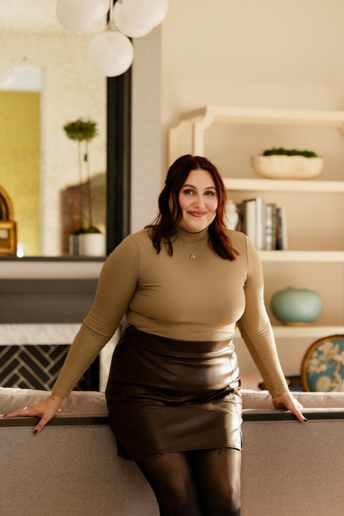 A woman in a tan top and brown skirt smiles while leaning against a sofa. The room has a neutral palette with bookshelves and plants.