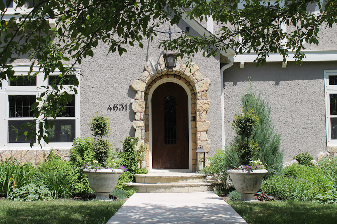 House entrance with arched wooden door, stone frame, and number 4631. Greenery and planters flank the walkway under leafy branches.