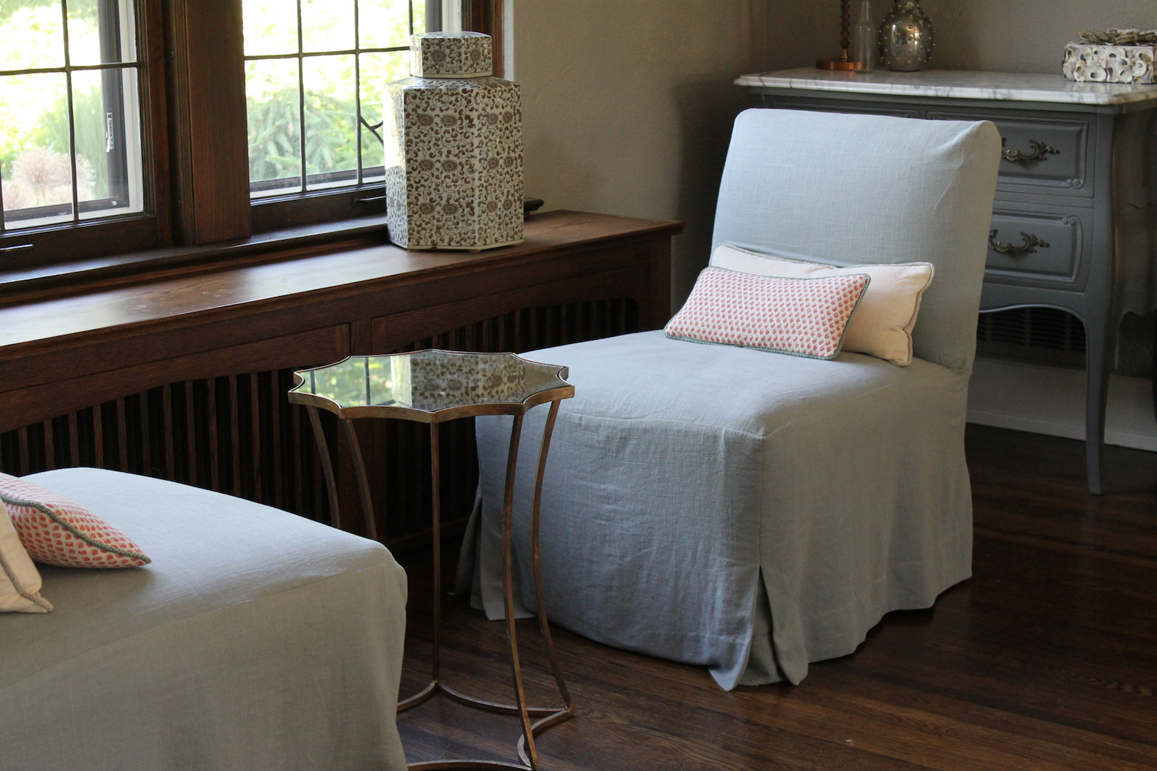 Cozy room with two gray chairs and patterned pillows. Elegant side table with a mirrored top. Wooden floor and window letting in natural light.
