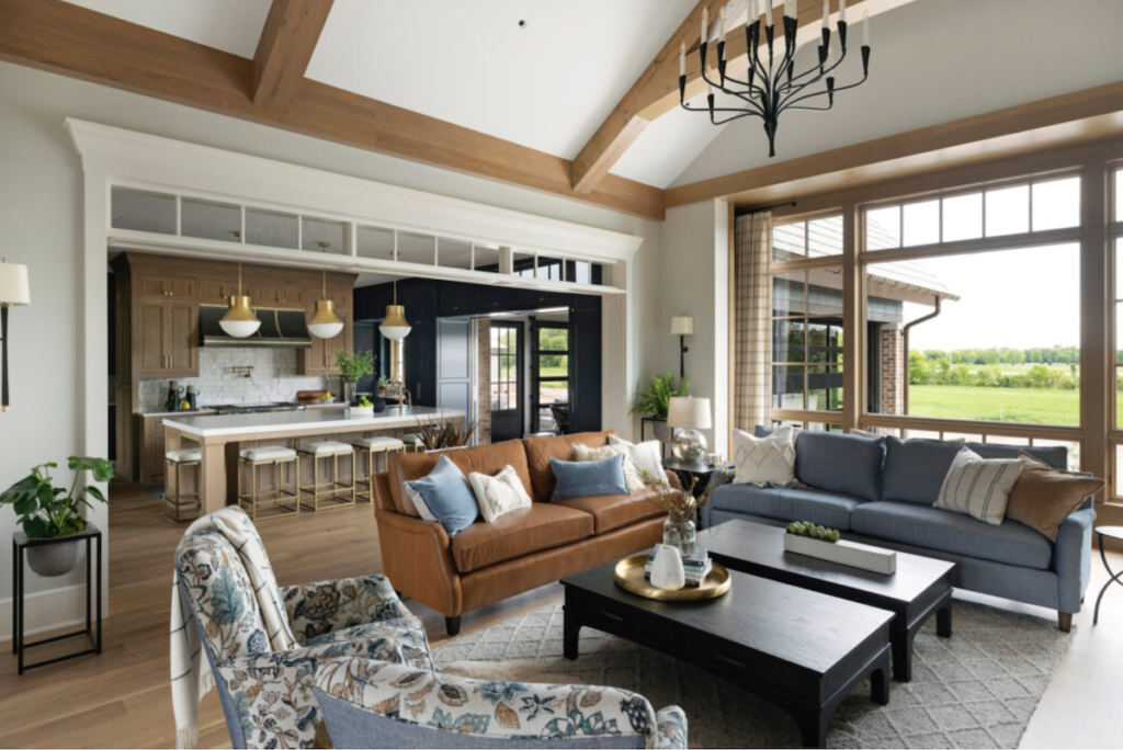 Spacious living room and kitchen with wooden beams, brown and blue sofas, floral armchair, large windows, and a modern hanging light.