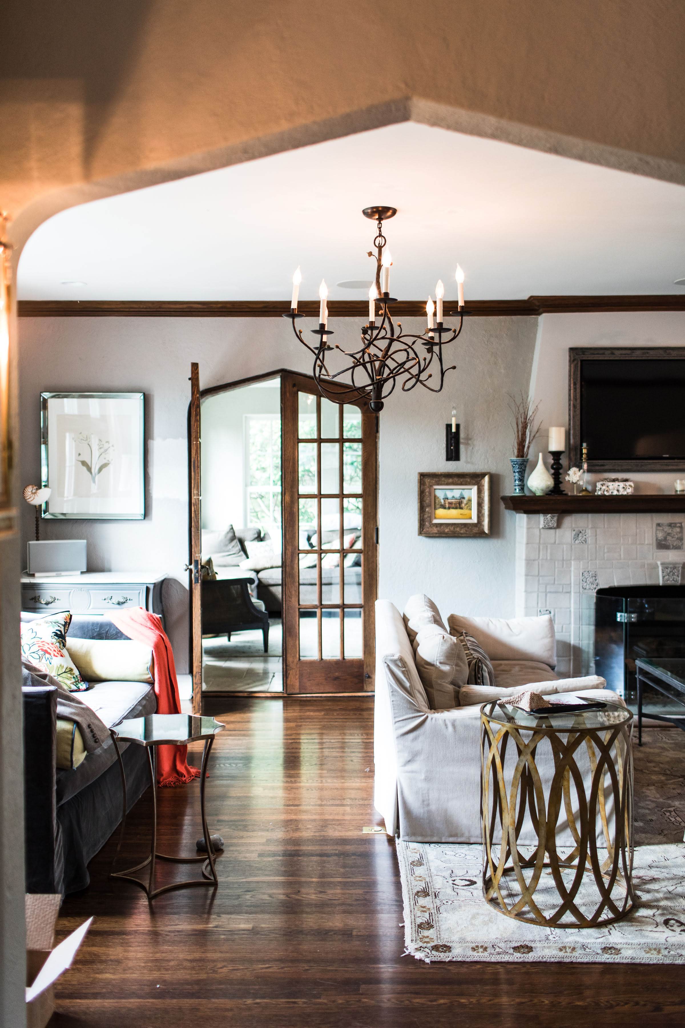 Cozy living room with beige sofas, elegant chandelier, and wooden floors. French doors open to sunlight; art and decor create a warm ambiance.