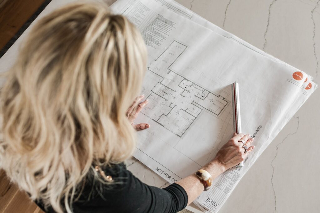 An interior designer examines architectural blueprints on a table, holding a scale ruler. The setting has a light, modern design.