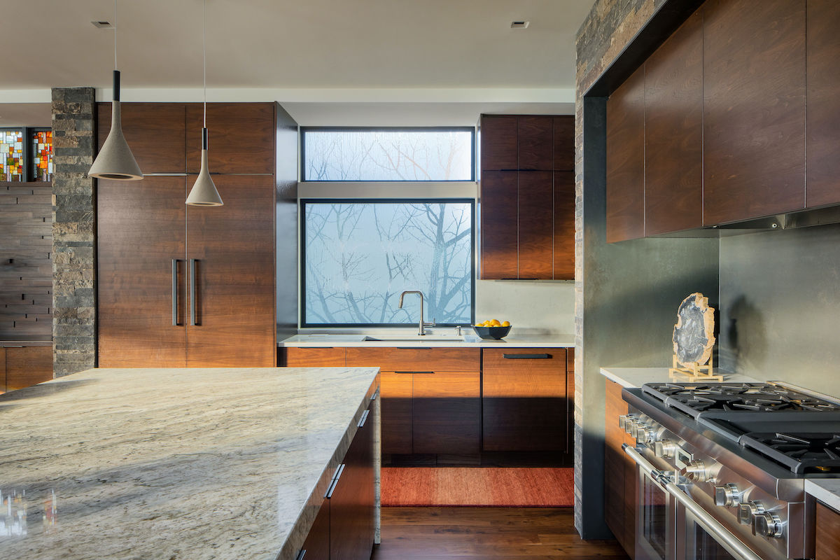 Modern kitchen with wooden cabinets, stone accents, and stainless steel stove. Large marble island and frosted window add elegance. Warm tones.